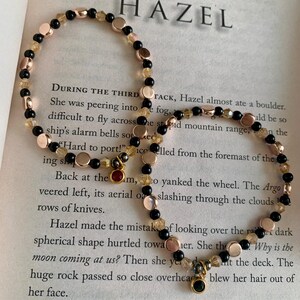 May include: Two beaded bracelets with the word "HAZEL" in the background. The bracelets feature black, gold, and rose gold beads. One bracelet has a small red charm, and the other has a green charm. The bracelets are on a book page.