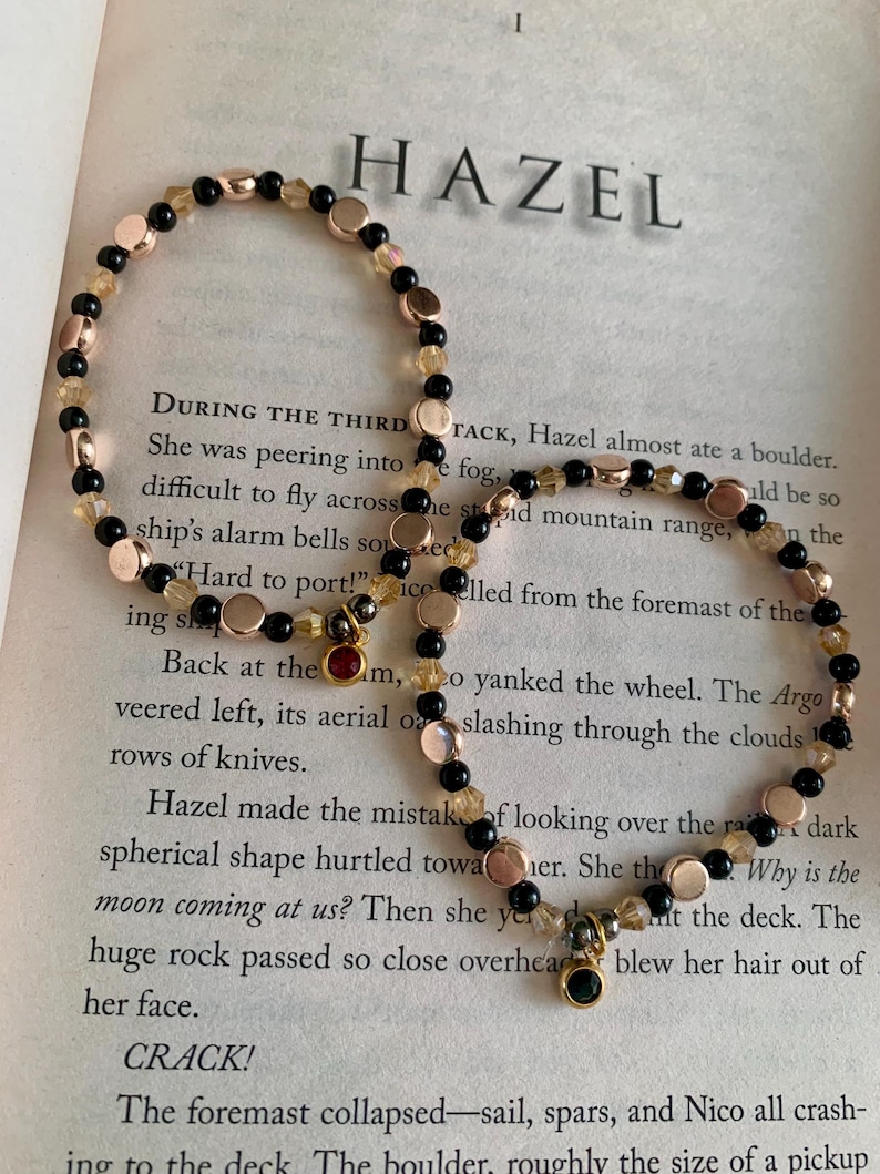 May include: Two beaded bracelets with the word "HAZEL" in the background. The bracelets feature black, gold, and rose gold beads. One bracelet has a small red charm, and the other has a green charm. The bracelets are on a book page.