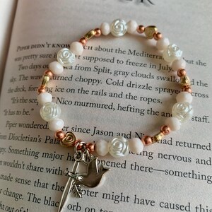 May include: A beaded bracelet with white rose-shaped beads, peach-colored beads, and gold-colored heart-shaped beads. A silver charm with a dove and a sword hangs from the bracelet. The bracelet is on an open book.