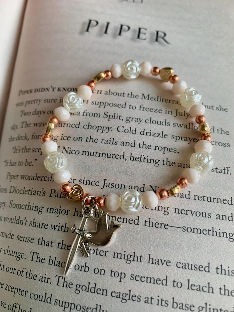 May include: A beaded bracelet with white rose-shaped beads, peach-colored beads, and gold-colored heart-shaped beads. A silver charm with a dove and a sword hangs from the bracelet. The bracelet is on an open book.