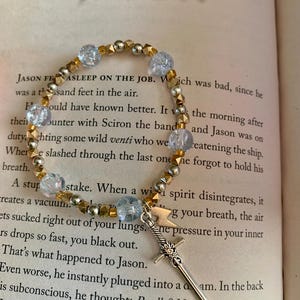 May include: A beaded bracelet with gold and silver beads, and light blue crystal beads. A silver sword charm hangs from the bracelet. The word "JASON" is visible in the background.