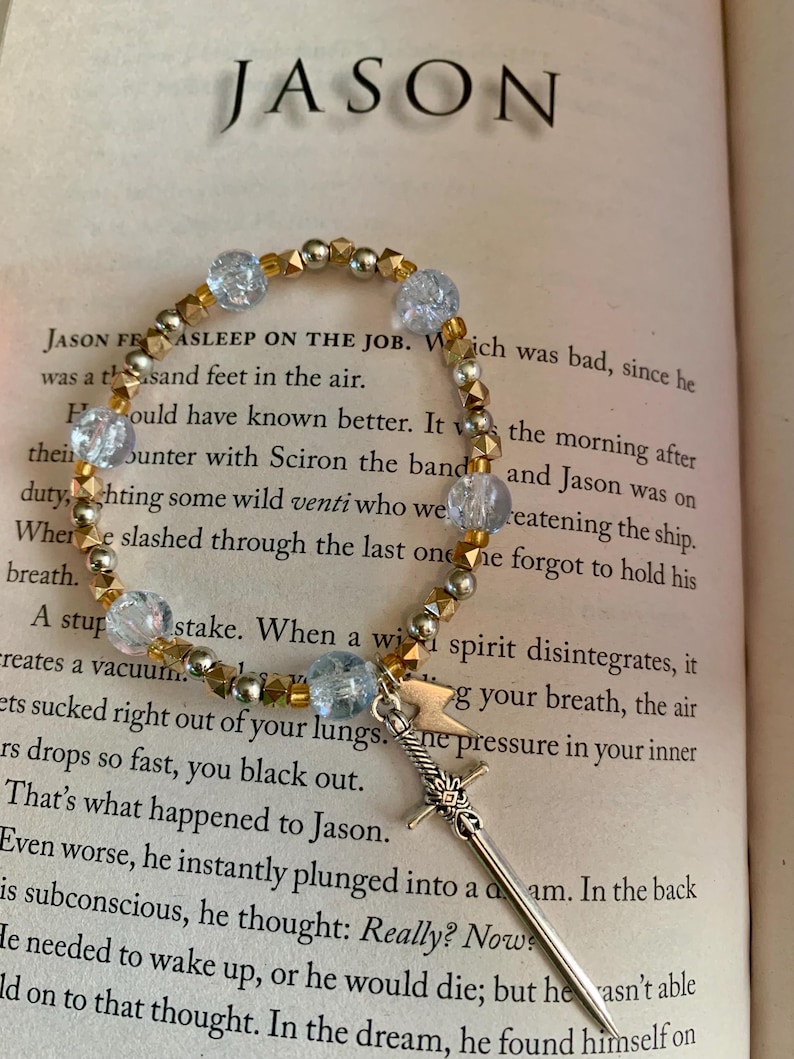 May include: A beaded bracelet with gold and silver beads, and light blue crystal beads. A silver sword charm hangs from the bracelet. The word "JASON" is visible in the background.