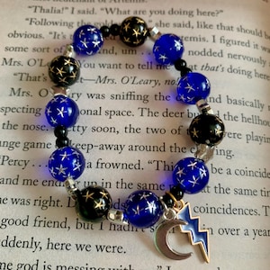 May include: A beaded bracelet featuring dark blue and black beads with star designs. The bracelet includes a silver charm with a crescent moon and a lightning bolt, set against a book page background.
