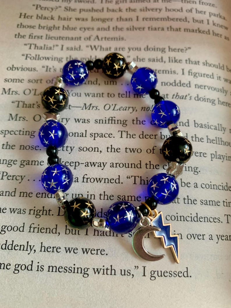 May include: A beaded bracelet featuring dark blue and black beads with star designs. The bracelet includes a silver charm with a crescent moon and a lightning bolt, set against a book page background.
