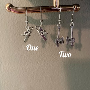 May include: Silver-toned metal earrings. One pair features chainsaw charms, and the other pair features axe charms. The word "One" is written above the chainsaw earrings, and the word "Two" is written above the axe earrings.