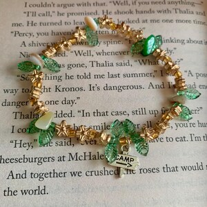 May include: A beaded bracelet featuring gold-colored star and bead accents, green leaf-shaped beads, and a small silver charm with the word "CAMP" and an arrow. The bracelet is on a background of open book pages.