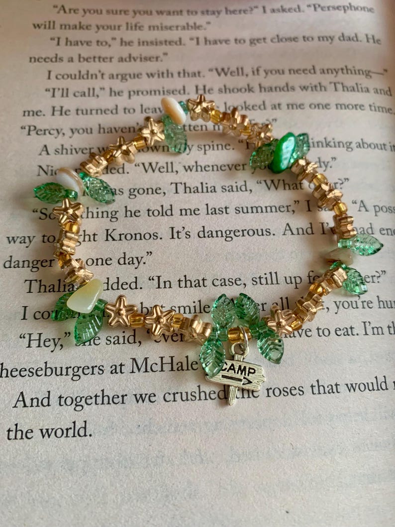 May include: A beaded bracelet featuring gold-colored star and bead accents, green leaf-shaped beads, and a small silver charm with the word "CAMP" and an arrow. The bracelet is on a background of open book pages.