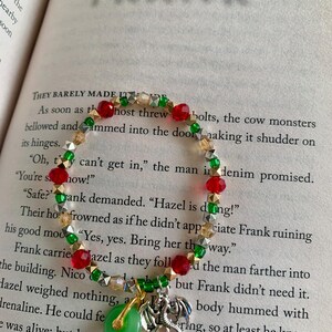 May include: A beaded bracelet with red, green, gold, and silver beads. The bracelet features a green jade pendant and a silver dragon charm. The word "FRANK" is visible in the background.