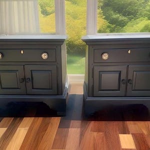 May include: Two dark blue nightstands with a single drawer and two doors. Each nightstand has a gold knob on the drawer and two gold knobs on the doors.