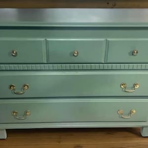 May include: A light blue dresser with four drawers and gold-toned handles. The dresser has a slightly distressed finish and sits on a wooden floor.