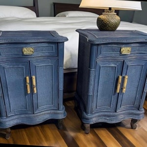 May include: Two navy blue bedside tables with woven door panels and bamboo-style handles. Each table has a drawer with a brass pull and sits on four curved legs. A lamp with a patterned base rests on one table.