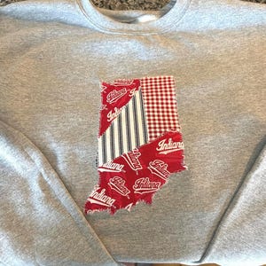 May include: A gray sweatshirt featuring a patchwork design of the state of Indiana. The Indiana shape is composed of red fabric with the word "Indiana" printed on it, along with red and white gingham and blue and white striped fabric.