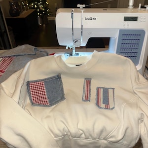 May include: Cream-colored cropped sweatshirt with three denim and red-and-white checkered patches. A Brother sewing machine is in the background, with a Christmas tree with lights in the distance. The sweatshirt is on a table.