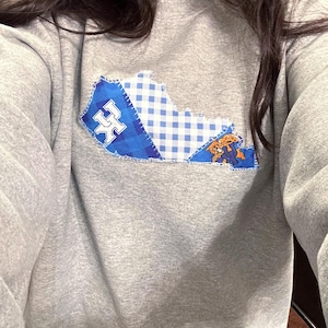 May include: A gray sweatshirt featuring a patchwork design of the state of Kentucky. The design incorporates blue plaid, a blue and white pattern, and a logo. The sweatshirt is a comfortable, casual garment.