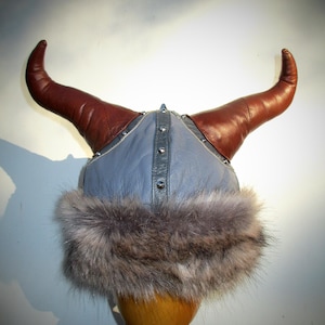 Puede incluir: Un gorro vikingo de cuero azul con cuernos de cuero marrón y un borde de piel gris.