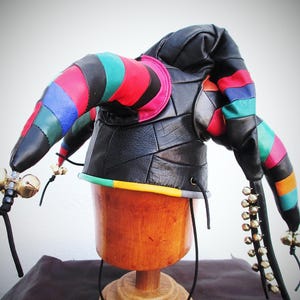 Könnte beinhalten: Ein schwarzer Leder-Narrenhut mit bunten gestreiften Hörnern und goldenen Glocken. Der Hut hat ein Patchwork-Design und eine grüne, gelbe und orangefarbene Bordüre. Die Hörner sind rot, blau, grün und schwarz gestreift.