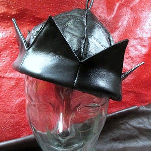 May include: A black leather hat with a crown-like design. The hat has a pointed shape with angular edges and a small loop at the top. The hat is displayed on a clear glass head.