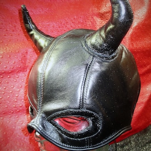 Puede incluir: Máscara de cuero negro con cuernos y agujeros para los ojos. La máscara está en una cabeza de maniquí.