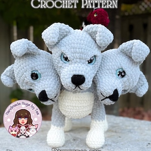 May include: A handmade crochet Cerberus dog with three heads, in shades of gray and white. The dog has blue eyes and black noses. The text "Prada The Cerberus Crochet Pattern" is at the top, and "Pattern by Handmade Hugs by Tea" is at the bottom.