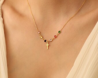 Puede incluir: Un collar de cadena dorada con un pequeño colgante de cruz y piedras preciosas de colores. El collar está en un modelo que lleva una blusa blanca.