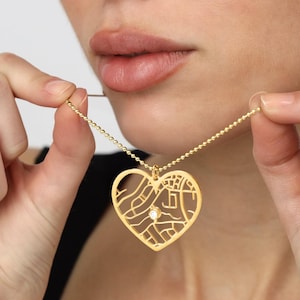 May include: A gold-colored heart-shaped pendant necklace with a map design and a small, clear gemstone. The necklace is held by a person, showcasing the delicate chain and pendant detail. The pendant's design features streets and buildings.