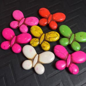 Op de afbeelding: Een verzameling vlindervormige kralen in verschillende kleuren, waaronder roze, rood, geel, groen en wit. De kralen hebben een glad, gepolijst uiterlijk en liggen op een donkere ondergrond.