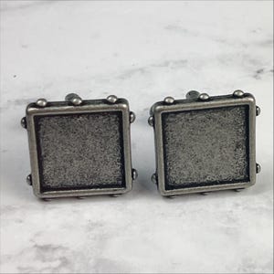 Puede incluir: Un par de gemelos de plata con un diseño cuadrado. Los gemelos tienen una superficie texturizada y un borde elevado.