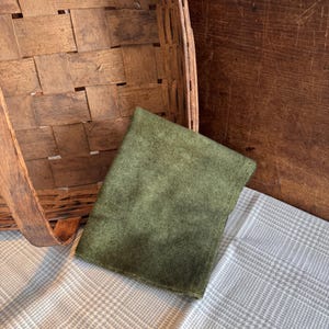 May include: An olive green fabric square rests on a gray and white plaid patterned cloth. A woven wooden basket and a wooden surface are in the background. The fabric square is a solid color and appears to be made of a soft material.