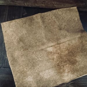 May include: A square, tan-colored piece of fabric with a slightly rough texture. The fabric has darker brown patches and appears to be folded. The background is a dark wooden surface.
