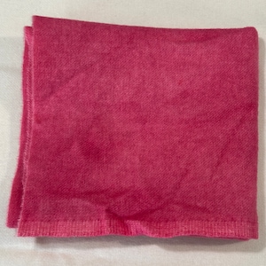 May include: A folded, square, magenta-colored cloth napkin. The fabric has a textured appearance, with visible weave details. The edges are neatly hemmed, and the overall presentation is clean and simple. The napkin is likely made of cotton or a similar material.