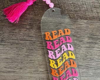 Peut inclure: Un marque-page en acrylique transparent avec un pompon rose et des perles roses. Le marque-page comporte le mot "READ" répété sept fois dans un arc-en-ciel de couleurs, y compris le rose, l'orange et le jaune.