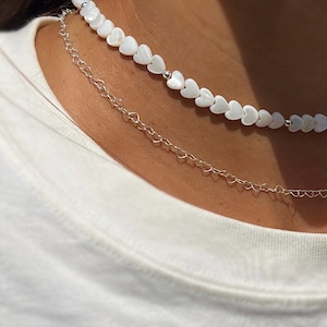 Può includere: Un girocollo con perline bianche a forma di cuore sovrapposto a una catena d'argento. Le collane sono indossate da una persona con una maglietta bianca.