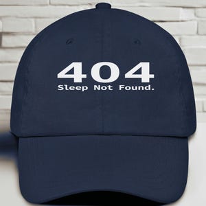 May include: Navy blue baseball cap with the text "404 Sleep Not Found" in white. The cap has a curved brim and a fabric strap closure. The hat is made of a durable material.