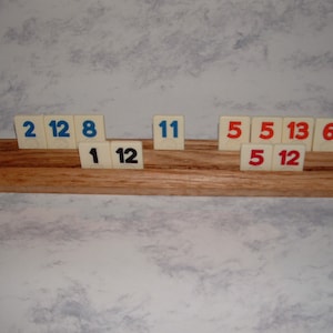 May include: A wooden rack with a set of white tiles with black and red numbers. The tiles are arranged in a row and display the numbers 2, 12, 8, 1, 12, 11, 5, 5, 13, 6, 5, and 12.