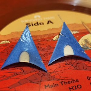 以下が含まれることがあります： 青と白の3Dプリントされた三角形のイヤリングが2つ。イヤリングは、赤いビニールレコードの上に置かれており、レコードには赤と白の火星風景のイラストが描かれている。レコードには「Side A」と「Running Time 20:35」の文字が書かれている。