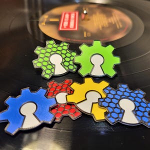 Könnte beinhalten: Sechs farbige Zahnrad-förmige Pins mit einem weißen Schlüsselloch-Design. Die Pins sind in einem Kreis auf einer schwarzen Oberfläche angeordnet.