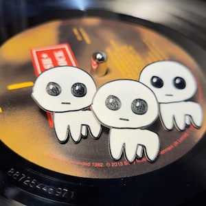 Puede incluir: Tres figuritas de cerámica blanca con ojos y bocas negras, de pie sobre un disco de vinilo negro. Las figuritas tienen forma de pequeñas personas redondas sin brazos ni piernas.