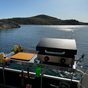 Puede incluir: Una parrilla portátil montada en un barco con una tabla de cortar y condimentos en una mesa lateral. La parrilla es negra con una base de acero inoxidable y dos perillas. El barco está en un lago con una montaña en el fondo.