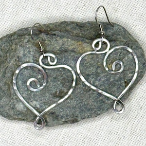 May include: A pair of silver heart-shaped earrings with a twisted wire design. The earrings are hanging from a silver hook.