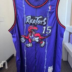 Puede incluir: Camiseta de baloncesto morada de los Raptors con ribete rojo y negro. Presenta el nombre del equipo "Raptors" en blanco, el número "15" y un logotipo de dinosaurio de dibujos animados. La camiseta tiene una textura de malla y un diseño de rayo.