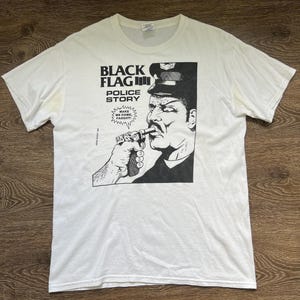Puede incluir: Una camiseta blanca de manga corta con un estampado gráfico negro en el pecho. El diseño incluye el texto BLACK FLAG, POLICE STORY y un bocadillo con el texto MAKE ME COME, FAGGOT! sobre una mano que sostiene una pistola hacia un policía.