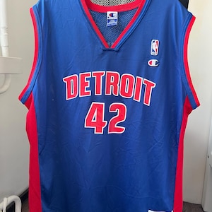 Puede incluir: Camiseta de baloncesto azul de Detroit con ribetes rojos y el número 42. La palabra "DETROIT" está impresa en el pecho en letras rojas. La camiseta tiene cuello en V y el logo de Champion.