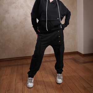 Peut inclure: Un sweat-shirt à capuche noir et un pantalon assorti avec un cordon de serrage et des détails boutonnés. Le mannequin porte des baskets blanches.