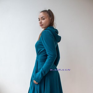 May include: A teal blue hooded dress with a long, flowing skirt and long sleeves. The dress has a high neckline and a fitted bodice. The model is wearing black boots.