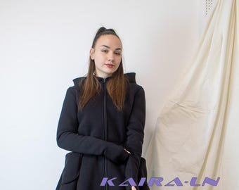 May include: A black hooded asymmetrical zip-up jacket with long sleeves and a high neckline. The jacket has a unique design with a long, flowing back panel and a shorter front panel. The model is wearing blue jeans and black knee-high boots.