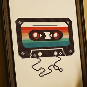 May include: A framed print of a pixelated cassette tape with rainbow colored stripes. The tape is in a brown frame and is hanging on a yellow wall.