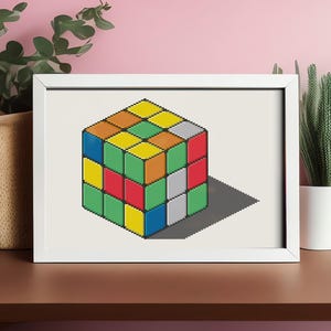 Op de afbeelding: Een wit ingelijste print van een kleurrijke Rubik's Cube met een schaduw eronder. De kubus is opgebouwd uit negen vierkanten aan elke kant, waarbij elk vierkant een andere kleur heeft: rood, oranje, geel, groen, blauw, wit en grijs.