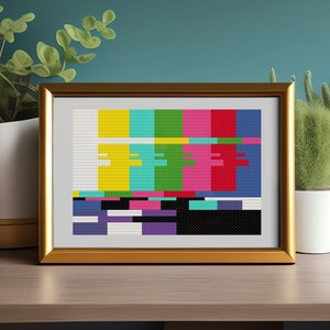 May include: A framed print of a pixelated television test pattern with a yellow, turquoise, green, pink, red, and blue colour scheme. The frame is gold and the print is on a wooden table with plants in pots.
