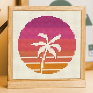 Puede incluir: Obra de arte de punto de cruz en un marco de madera. El diseño presenta una puesta de sol con un degradado de colores de naranja a rosa y morado. Una silueta de palmera blanca está en el centro.