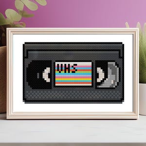 Puede incluir: Un patrón de punto de cruz de pixel art de una cinta VHS. La cinta es negra con una etiqueta de colores arcoíris que dice "VHS".